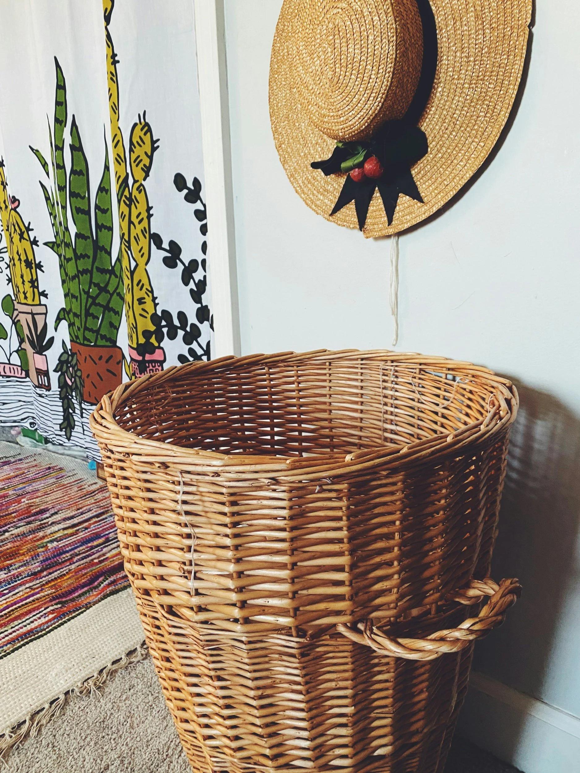 Empty woven wicker laundry basket in a room with a colorful rug, a wall hanging with cactus illustrations, and a straw hat with decorative berries hanging on the wall.