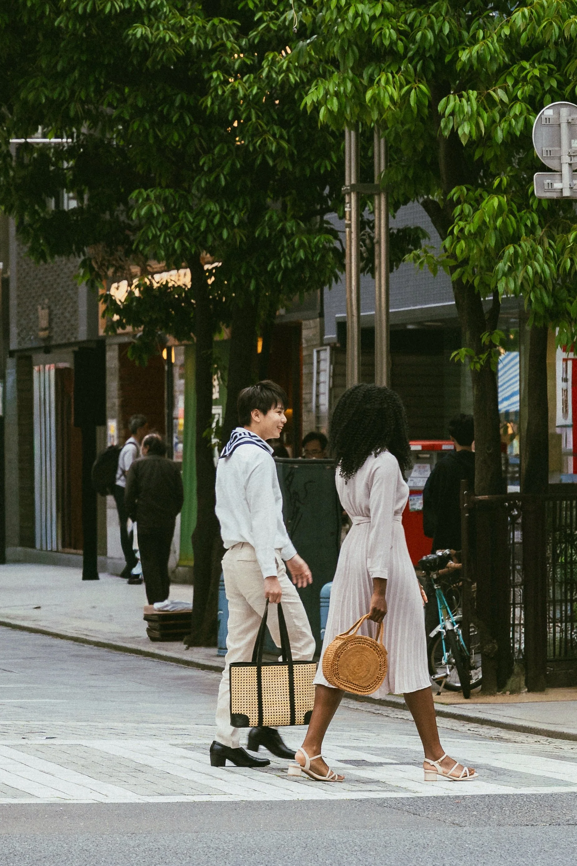 People walking on a city sidewalk with trees, buildings, and utility poles in the background.