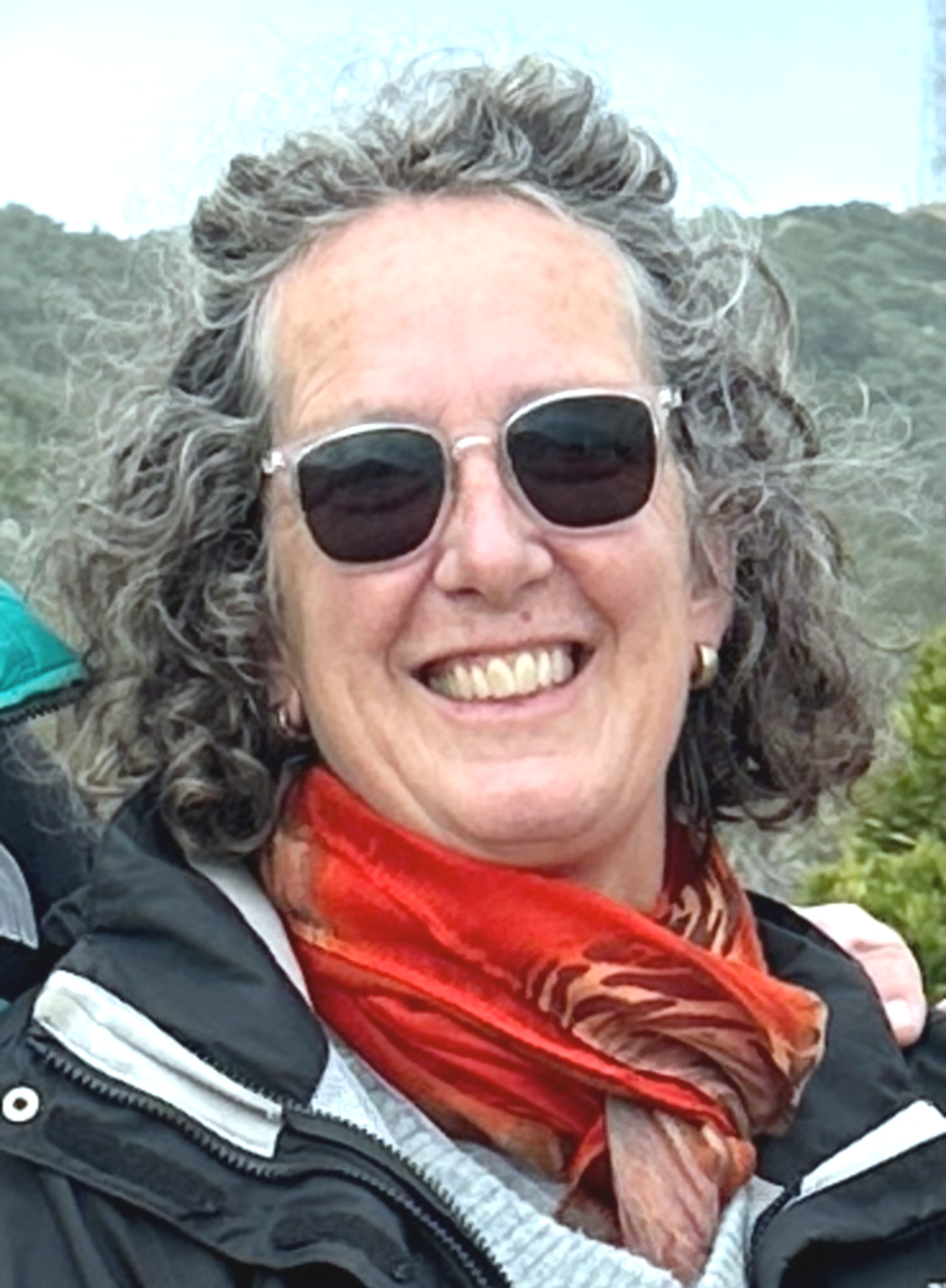 A woman with curly gray hair wearing sunglasses, a black jacket, and a red scarf, smiling outdoors.