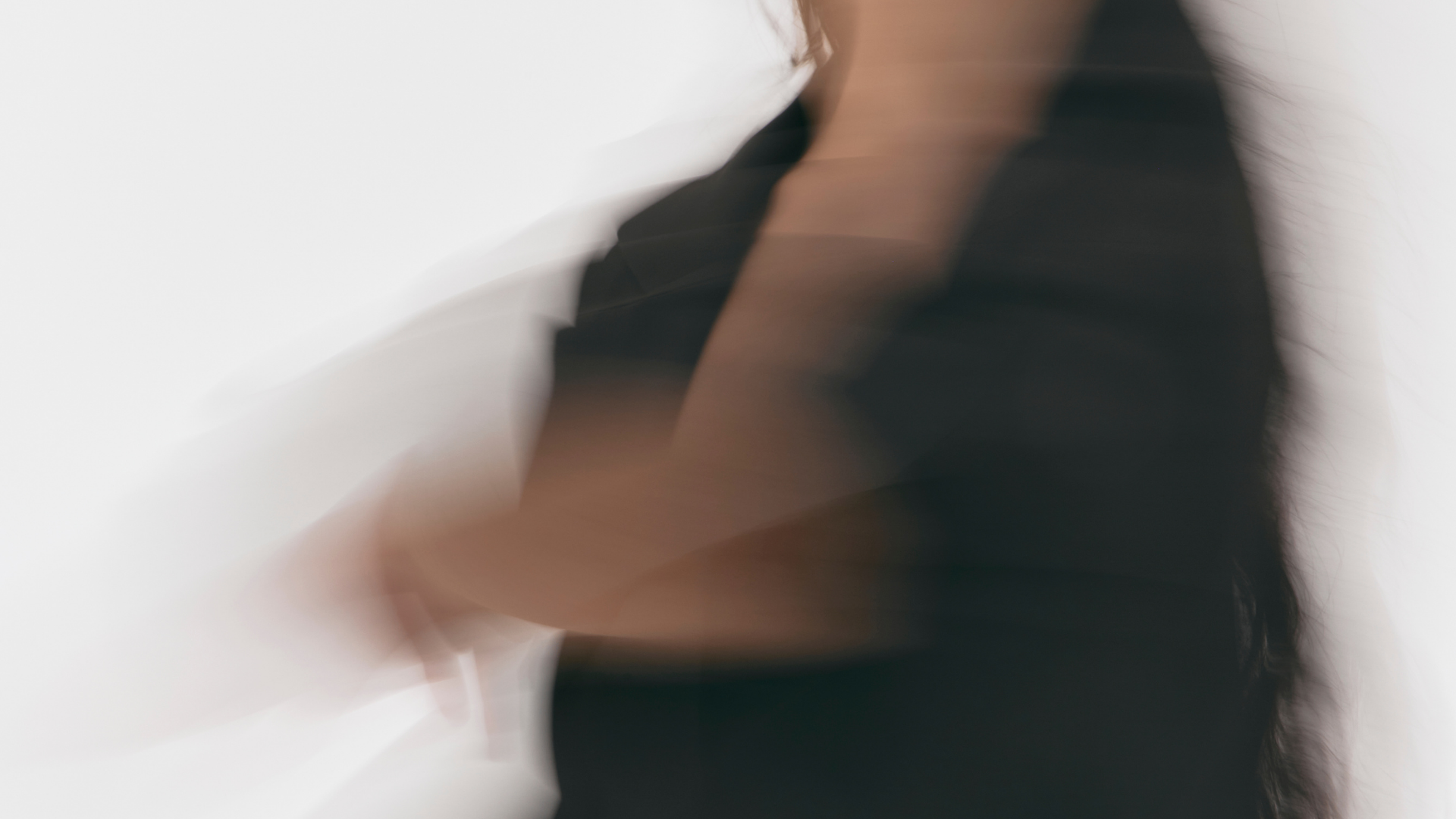 Blurred image of a woman with long dark hair, wearing a black top, standing against a light background.