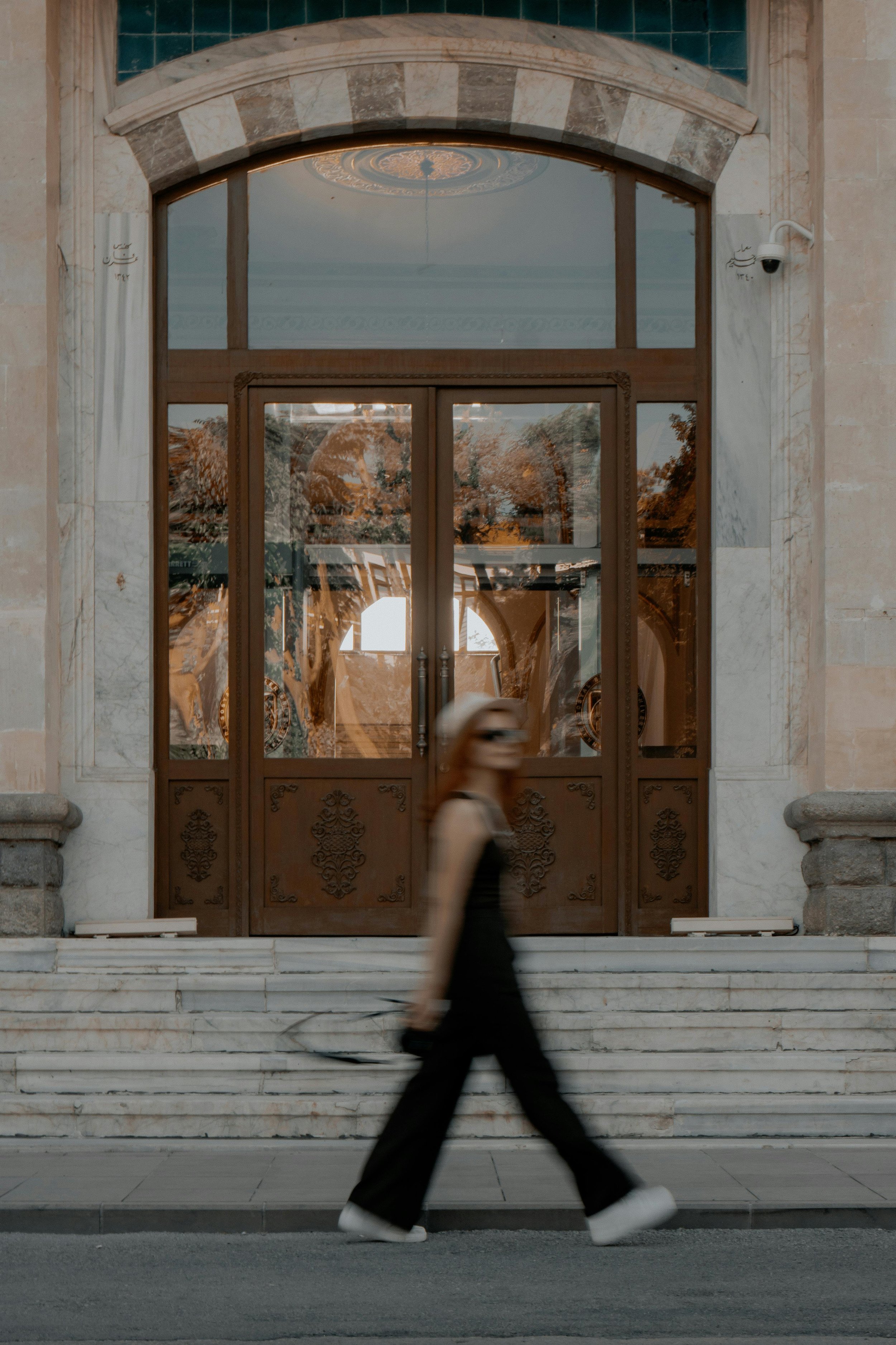 A woman in black clothing with white shoes and a white hat walking past an ornate wooden door with glass panels, set in a stone building with marble accents and stone steps.