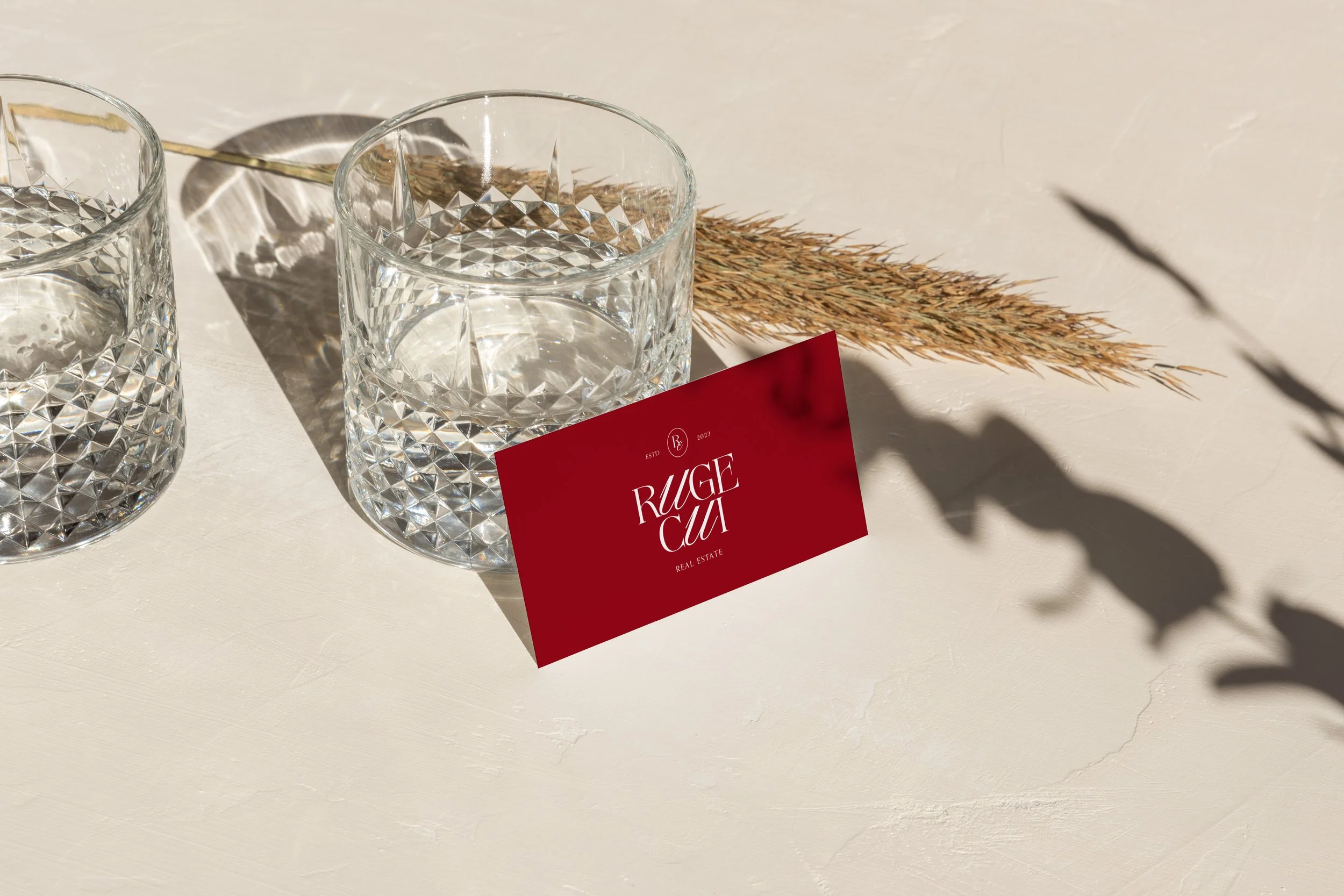 Two clear textured glasses filled with water, a red business card with white text reading 'RUGE CUT REAL ESTATE,' a dried plant stem, and shadows cast on a cream-colored surface.