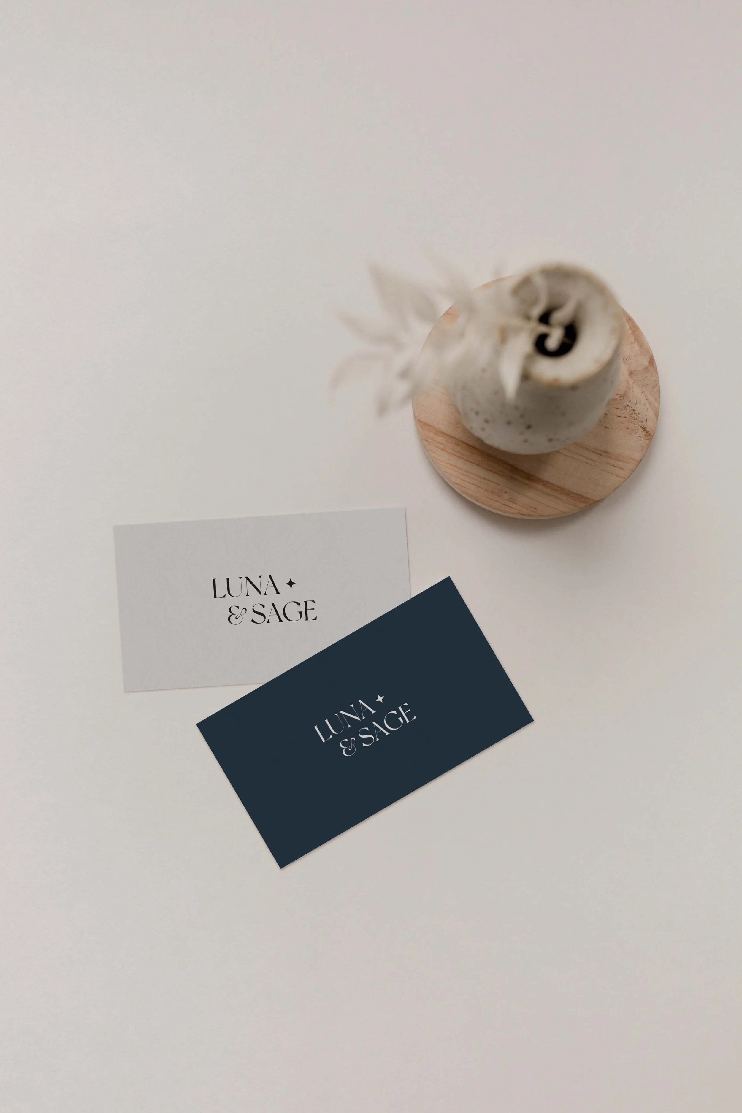 White business card and navy blue business card with 'LUNA & SAGE' text, placed on a white surface. A small white ceramic vase with a plant is positioned above the cards.