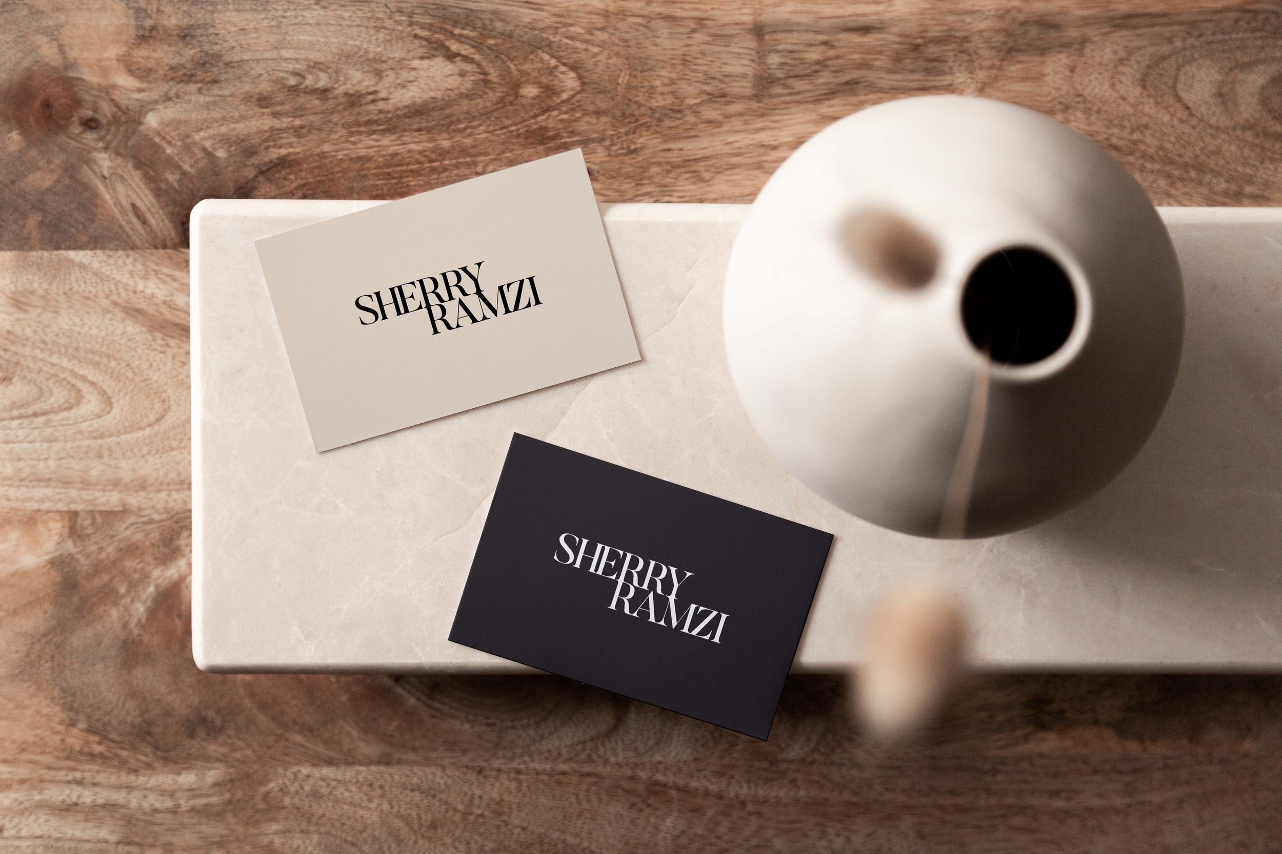 Two cards with the name 'SHERRY RAMZI,' one in white background with black text and the other in black background with white text, placed on a beige marble surface next to a white vase on a wooden surface.