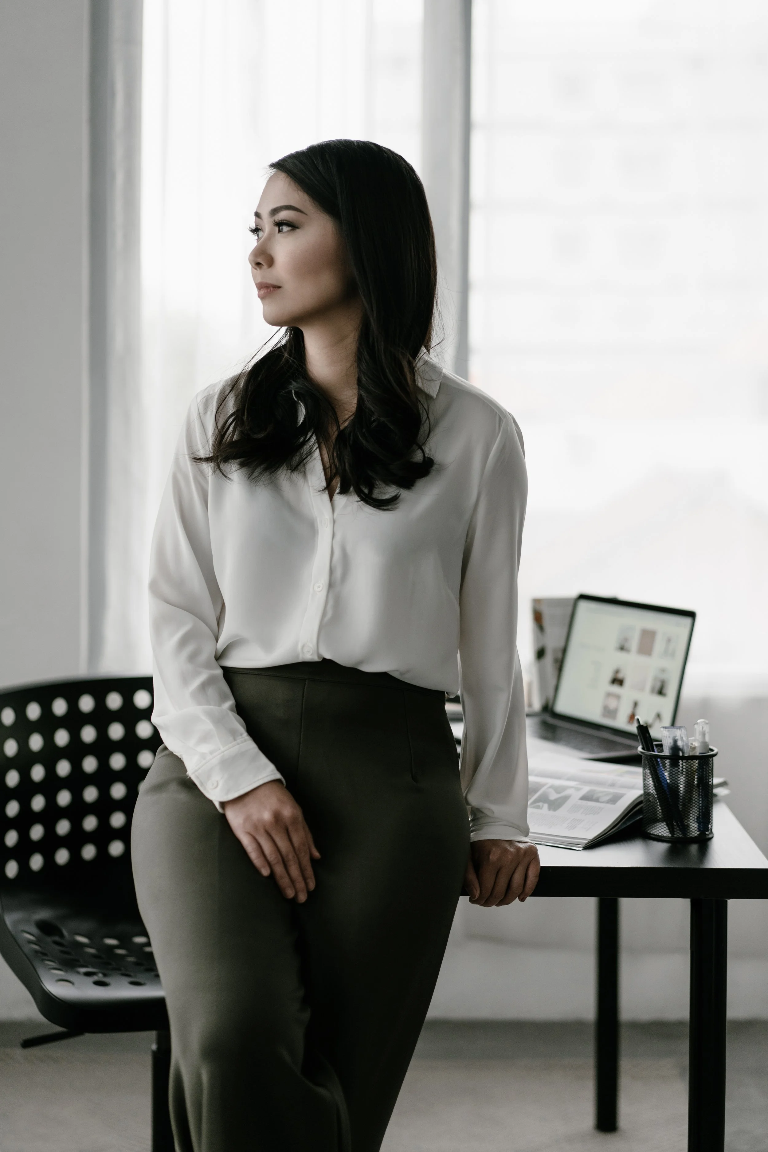A woman stands in an office, wearing a white blouse and gray pants, near a desk with a laptop and office supplies, looking out of the window to her left.
