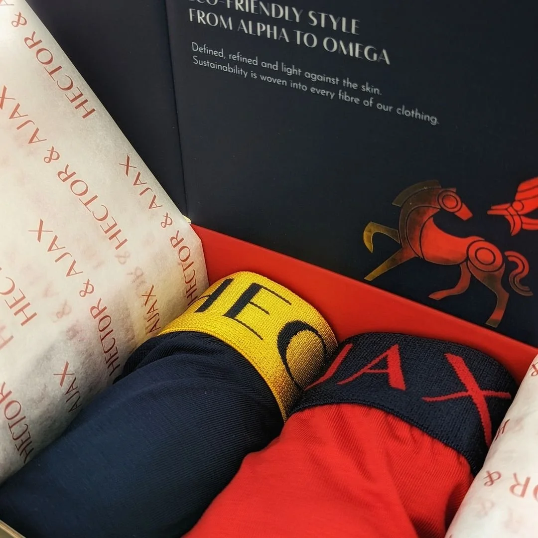 Close-up of two rolled-up socks, one in yellow with black text and the other in red with black and red text, inside a shoebox with tissue paper. The lid of the shoebox is dark with white text and a gold and red horse logo.