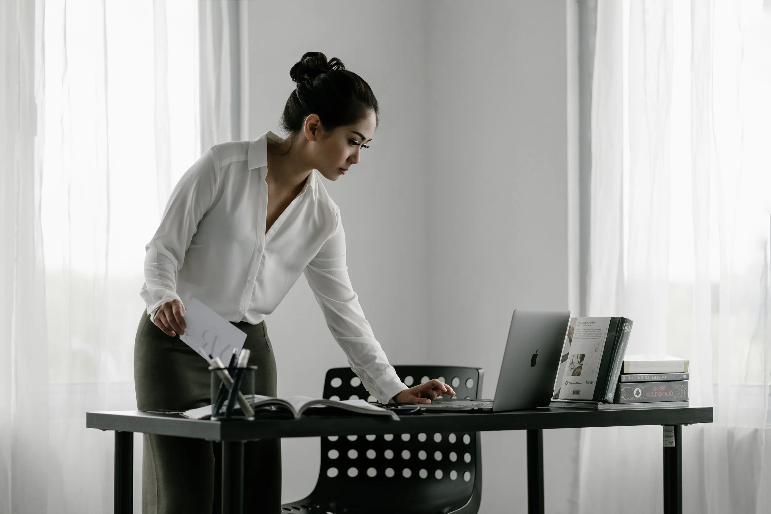 A woman in a white blouse standing at a desk, looking at a laptop with white curtains in the background.