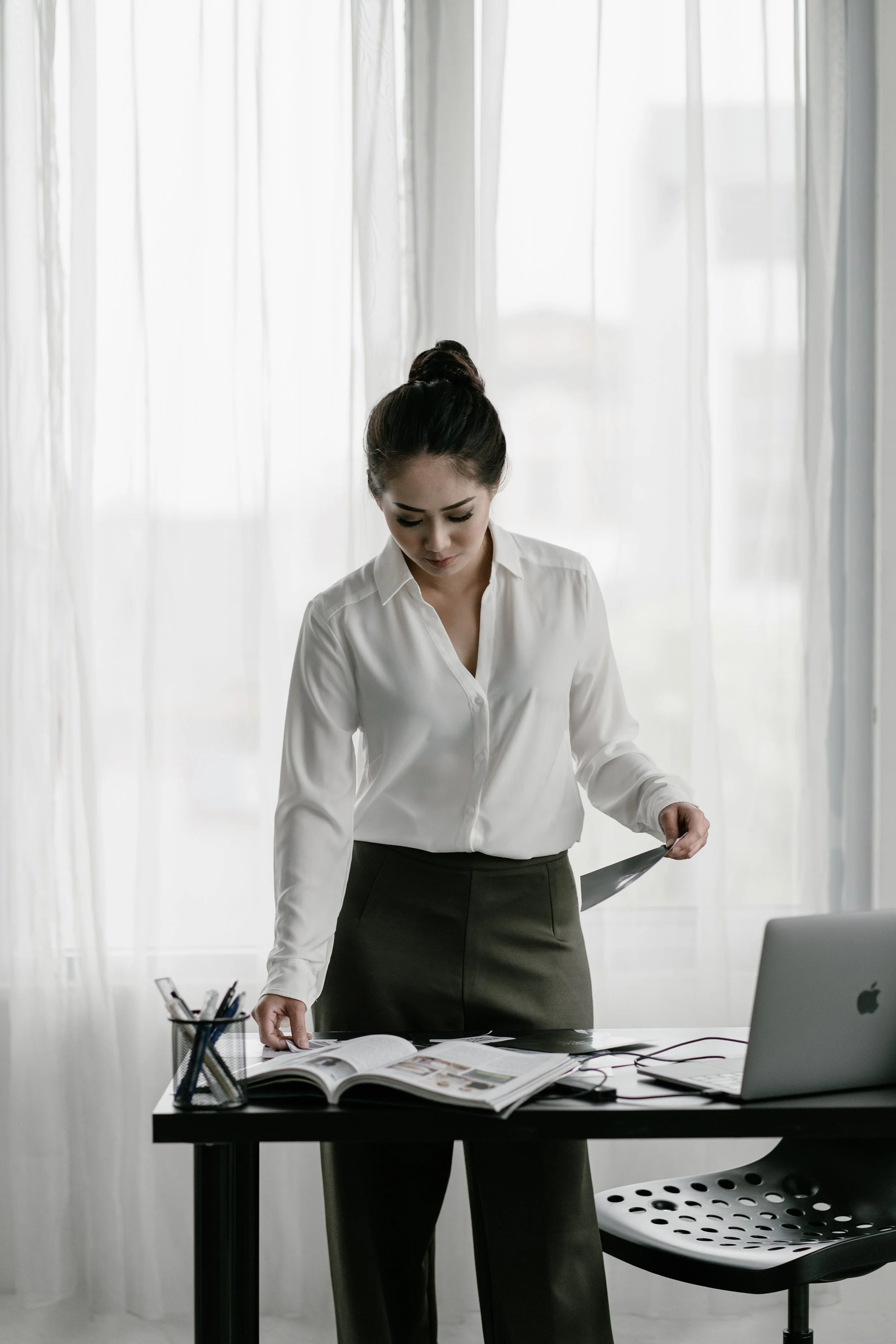A woman in a white blouse and dark pants standing at a desk, looking at open magazines, with a laptop and office supplies on the desk.