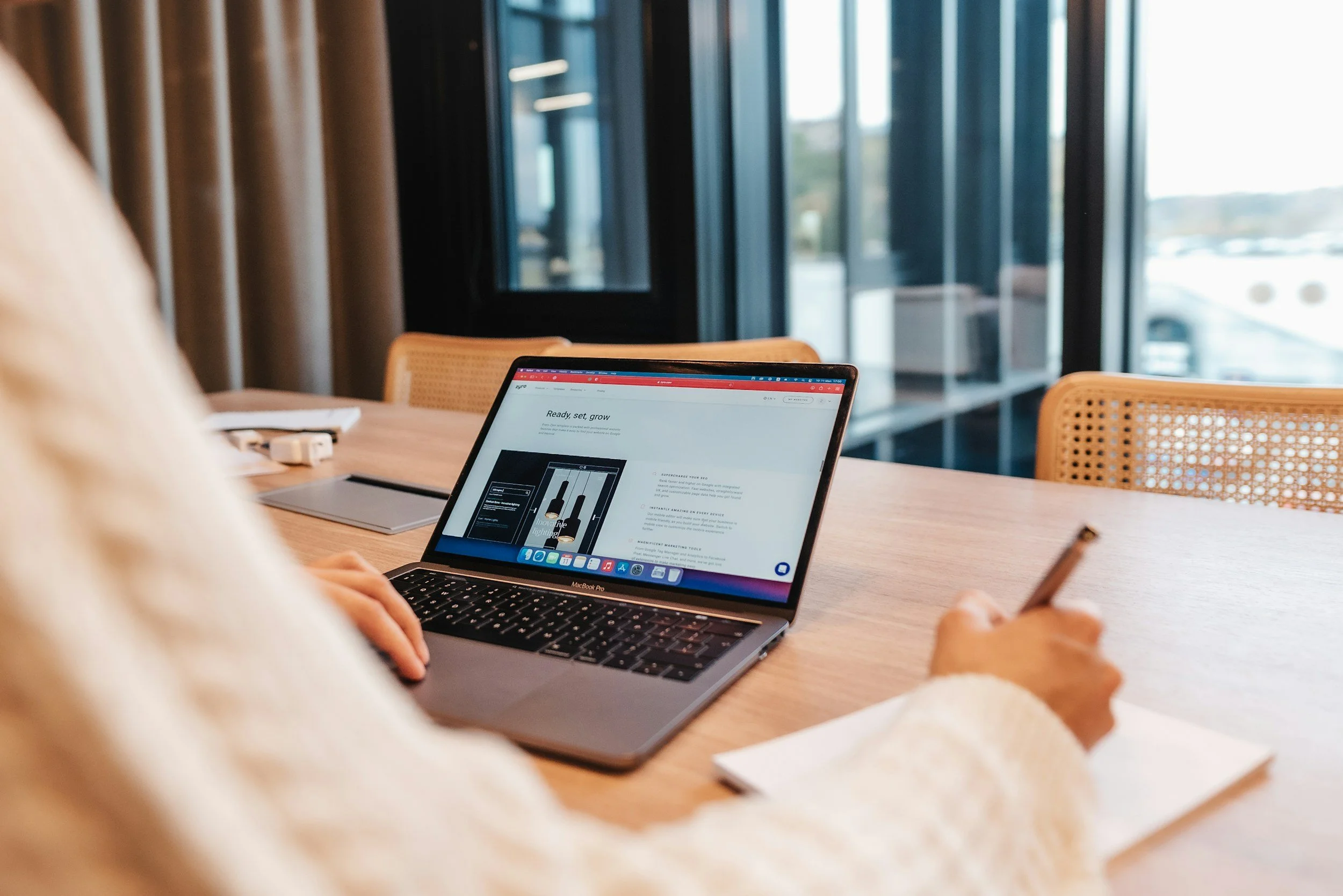 Person using a MacBook Pro laptop at a wooden conference table with a notepad and pen, in a modern office or cafe with large windows and natural light.