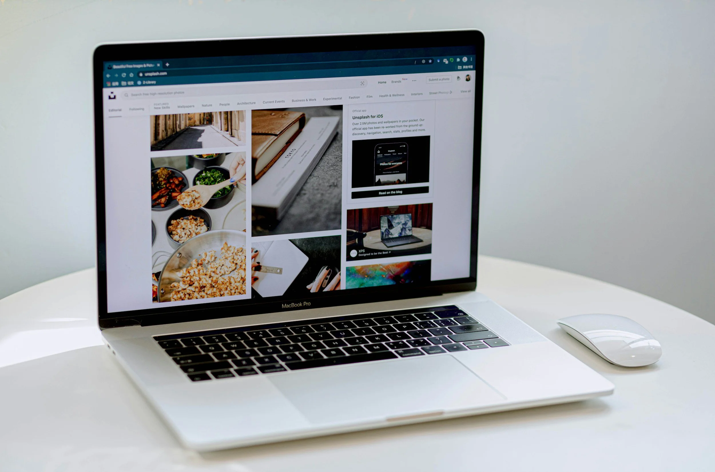 A MacBook Pro laptop displaying a webpage with images of food, books, and electronic devices on a white table, with a wireless mouse beside it.