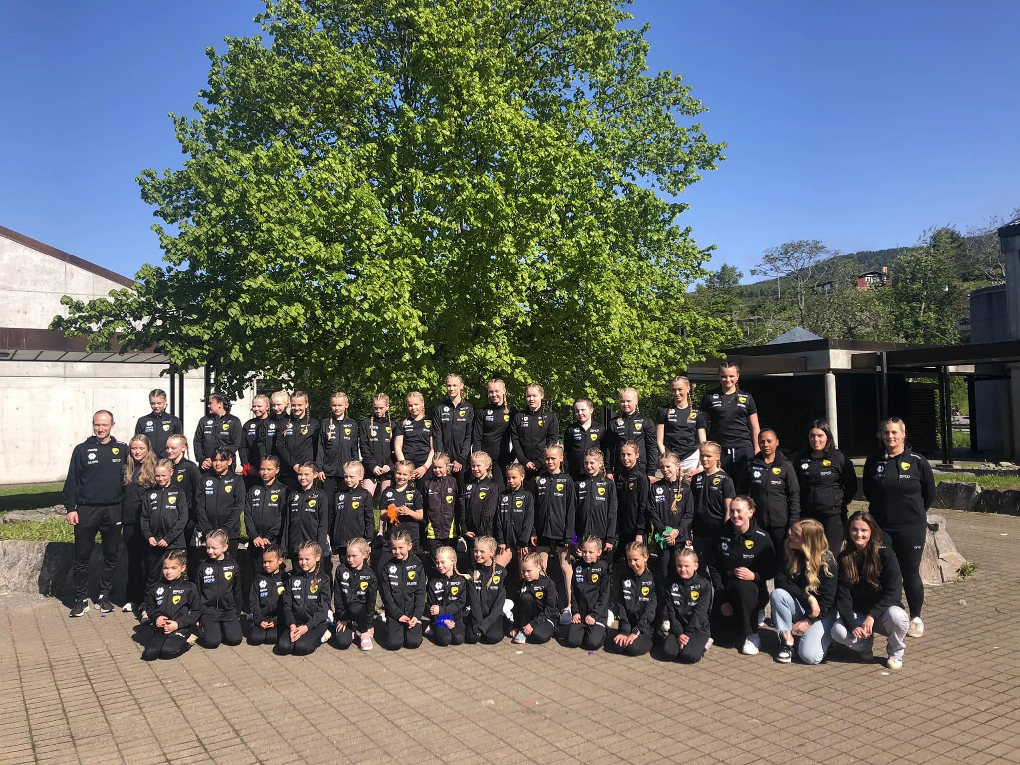 Ei stor gruppe med unge turnarar og trenarar står oppstilt utendørs, framfor eit stort tre
