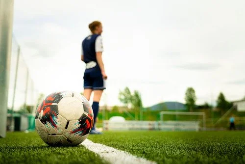 Bilde av en fotballbane. En fotball er i fokus, og i bakgrunnen ser man en fotballspiller med ryggen til kamera og en fotballbane