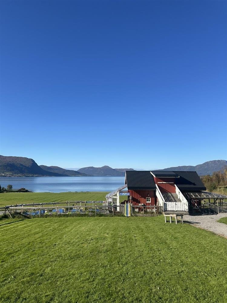 Grønt gresslette med et lite, rødt hus ved vannet, omgitt av fjell under en klar blå himmel.