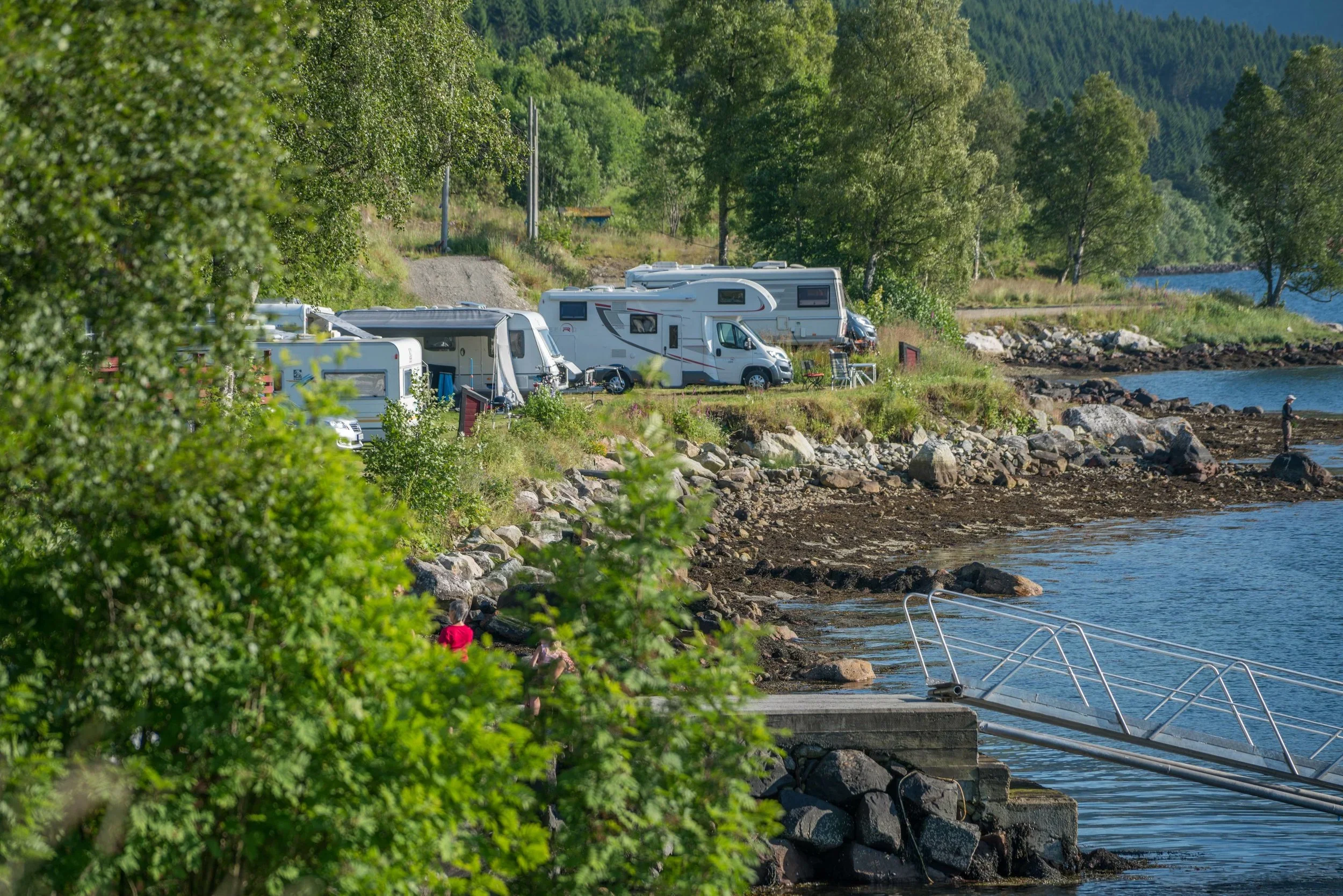 Campingvogner parkert ved sjøen omringet av grønne trær med fjell i bakgrunnen.