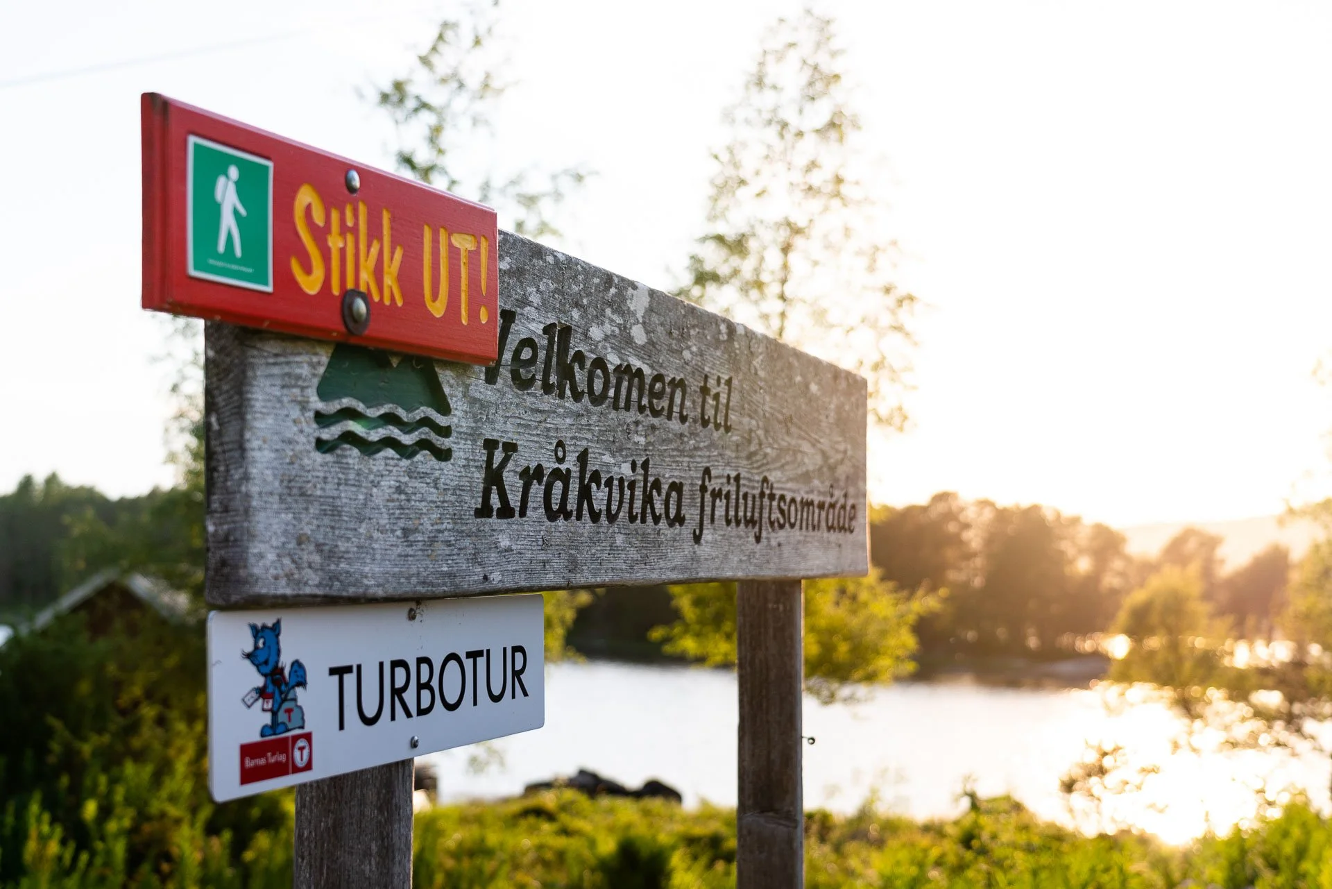 En trestolpe med tre skilt: rød med gul tekst som sier 'Stikk ut!', et treskilt med teksten 'Velkommen til Kråkvika friluftsområde', og en hvit med en blå og rød maskot og teksten 'Turbotur'. Vann og skog i bakgrunnen.
