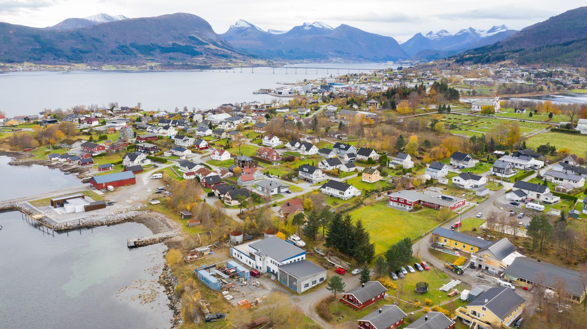 Dronebilde av Vestnes. Liten by ved fjord med fjell i bakgrunnen, små hus, båter, og veier.