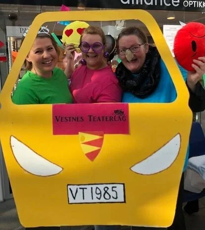 Tre kvinner er utkledd med artige briller, hodepynt og leikenase. Dei står med ei gul bilderamme som forestiller ein buss eller bil, og med teksten vestnes teaterlag frampå bilen