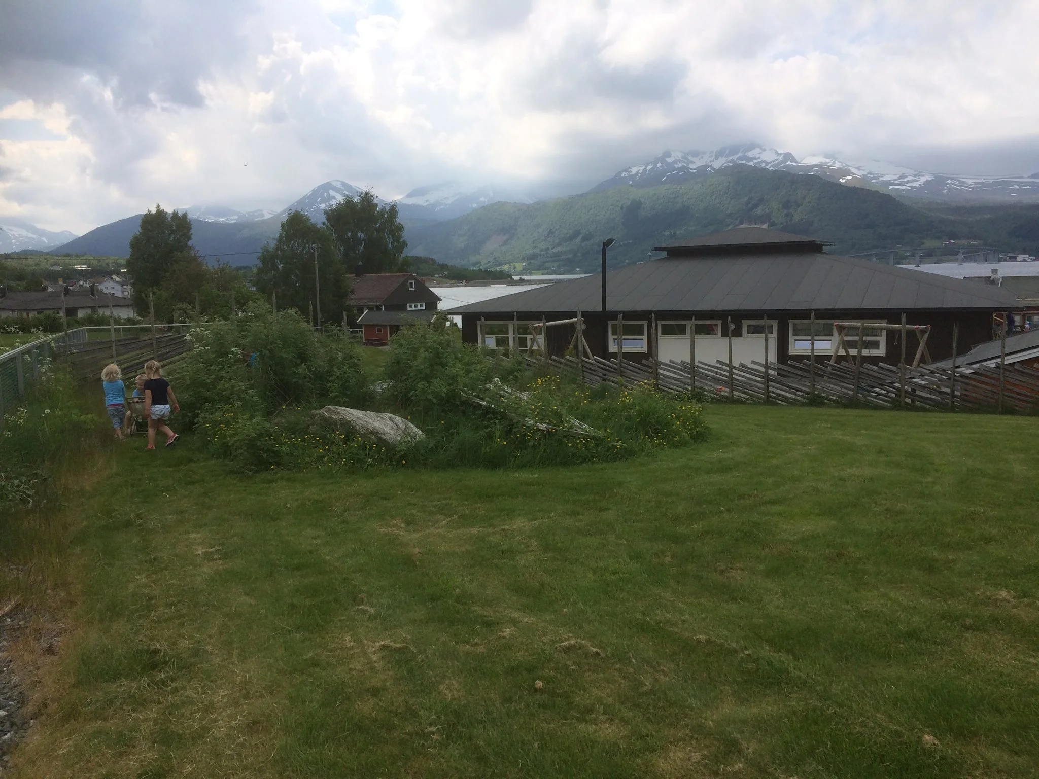 To barn leker i en grønn hage med trær og store steiner, omgitt av landskap med fjell og skyer i bakgrunnen.