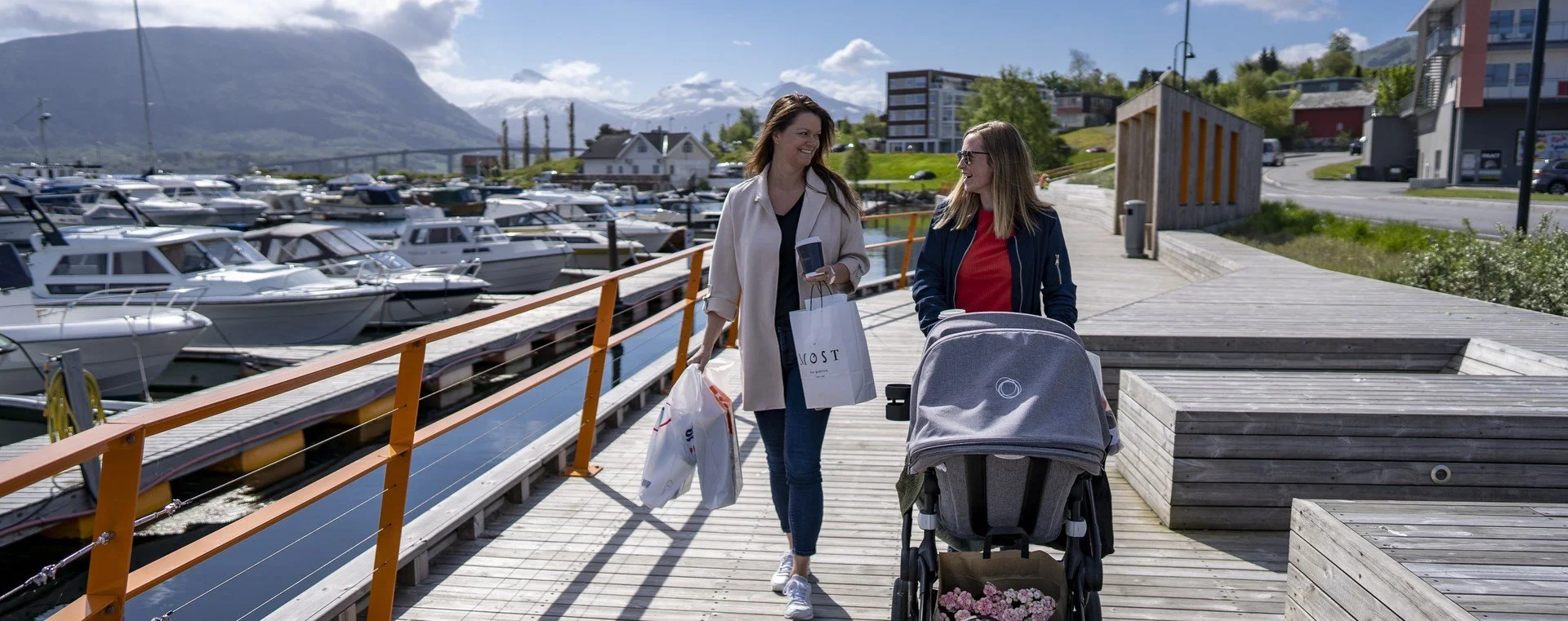 To kvinner går langs en båthavn, en av dem med en barnevogn og handleposer. De snakkar og smiler.