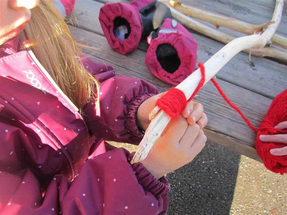 Barn som holder en pinne med rød garn, på et trebord med garn i hatter i bakgrunnen.