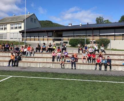 Barn og voksne som sitter på tribunen fremfor fotballbanen ved skolen, en solrik dag.