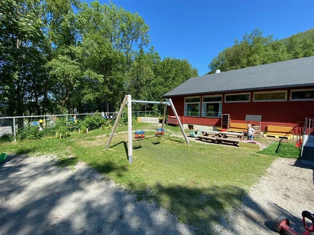 Uteområde med hus, lekeplass, huske og benker, omgitt av grønne trær på en klar dag.