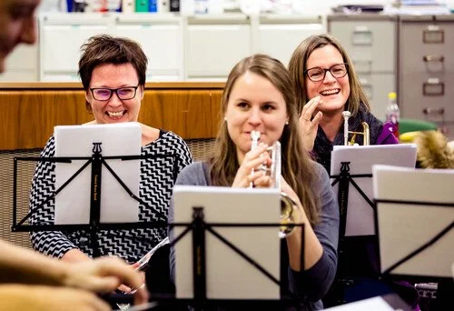 Tre damer på ulike instrument smiler mot kamera. Dei har notestativ framfor seg og held på med ei korpsøving