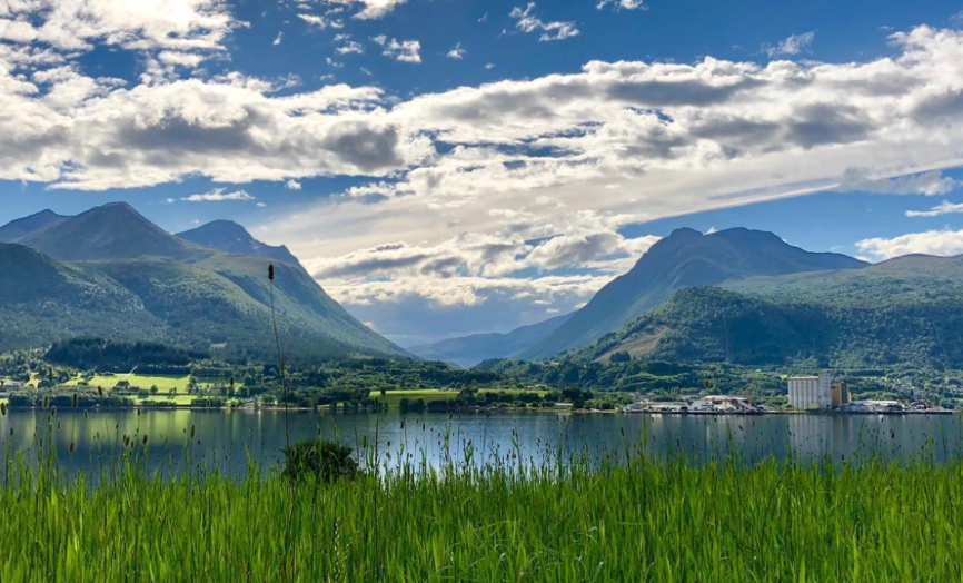 I forgrunnen en grønn plen. I bakgrunnen stille hav, fjell og en lettskyet himmel.
