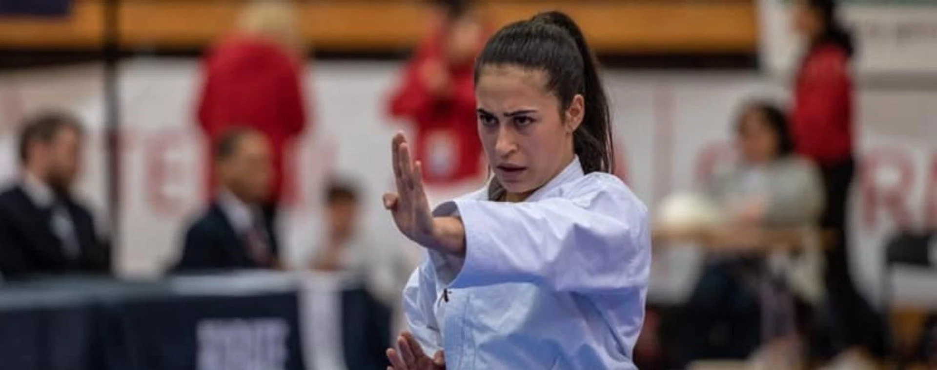 Ei kvinne utøver kampsport. Ho har på seg ei kvit drakt og gjennomfører ein øvelse i ein hall. Ho har eit bestemt og fokusert blikk, ned ei hand retta opp framfor seg