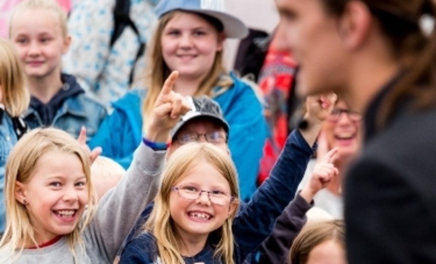 En gruppe glade barn som smiler og peker mot en voksen person, utendørs.