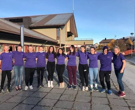 En gruppe ungdommer står side om side utendørs, fremfor skolen, alle iført matchende lilla t-skjorter.