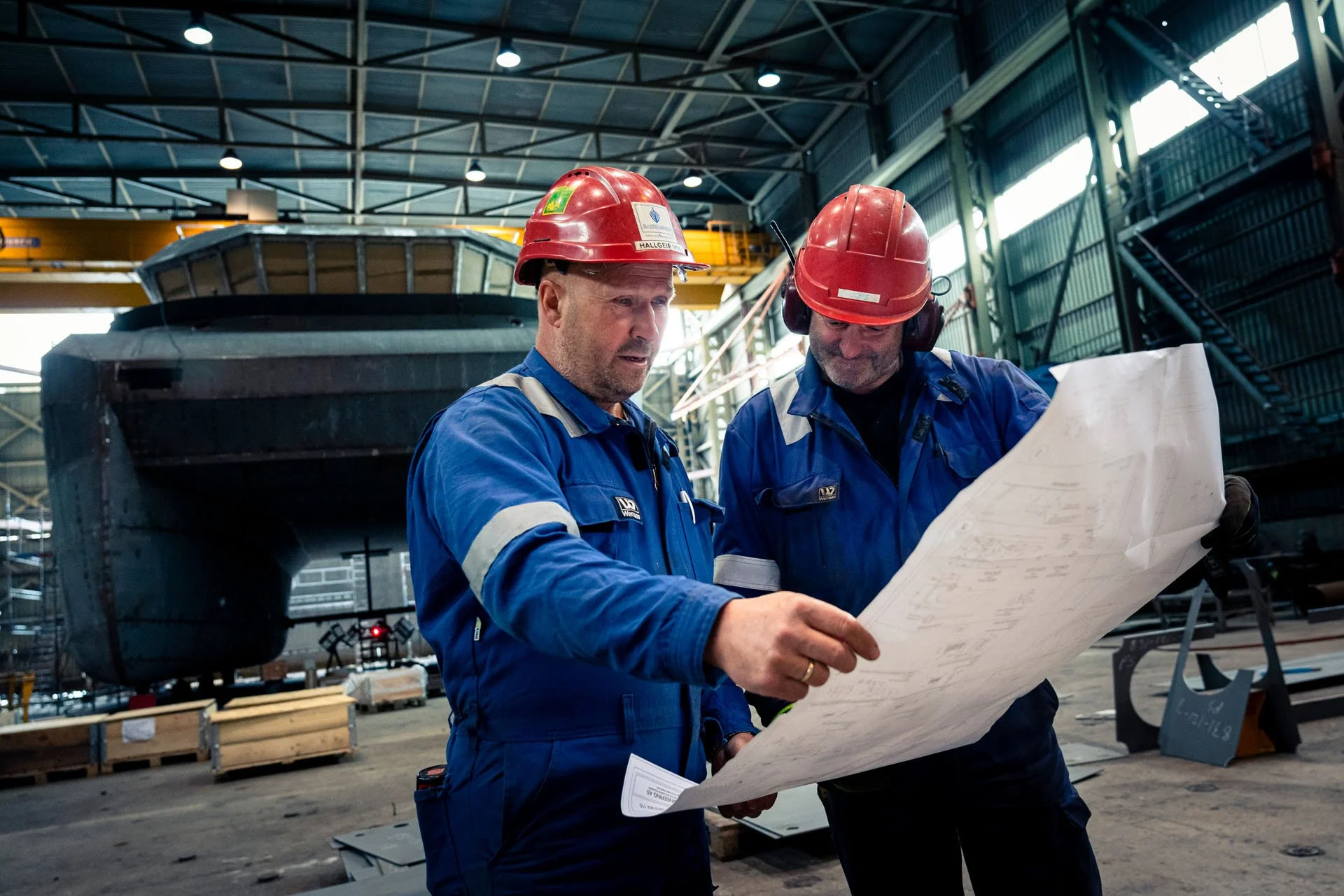 To menn i blå kjeledress og rød hjelm som ser på en teknisk tegning foran en stor båt i et verksted.