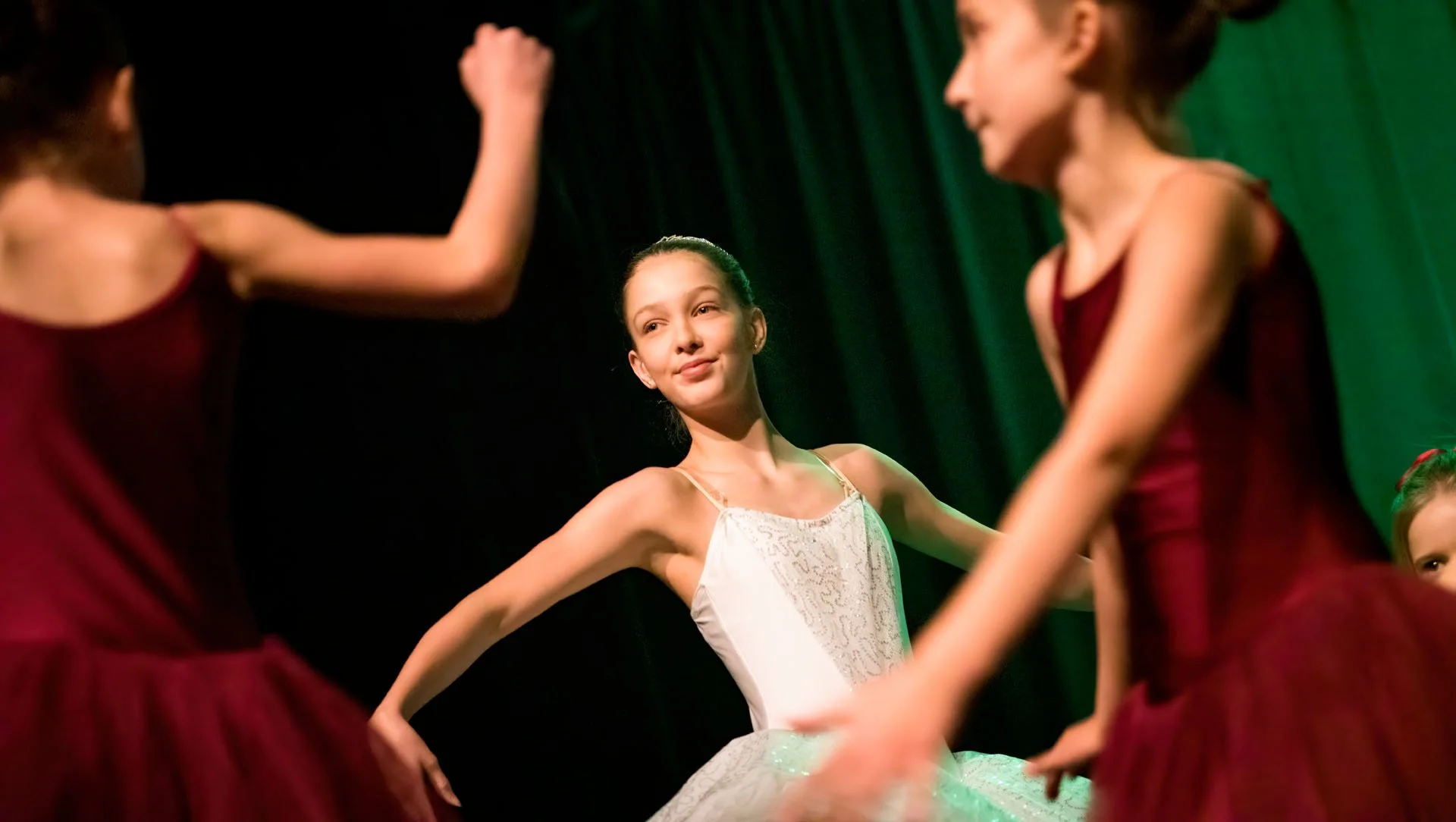 Ei ung jente i kvit kjole som dansar ballett er i fokus. To andre unge jenter i mørk raud kjole dansar rundt ho