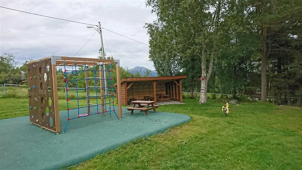 En lekeplass med klatrevegg og nett, en trehytte med piknikbord, gresslette, og trær i bakgrunnen.