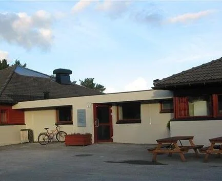 En enkel, hvitmalt bygning med en rød inngangsdør og vinduer, en sykkel parkert mot veggen, og en utendørs benk eller piknikbord foran bygningen, med klar himmel og noen skyer i bakgrunnen.