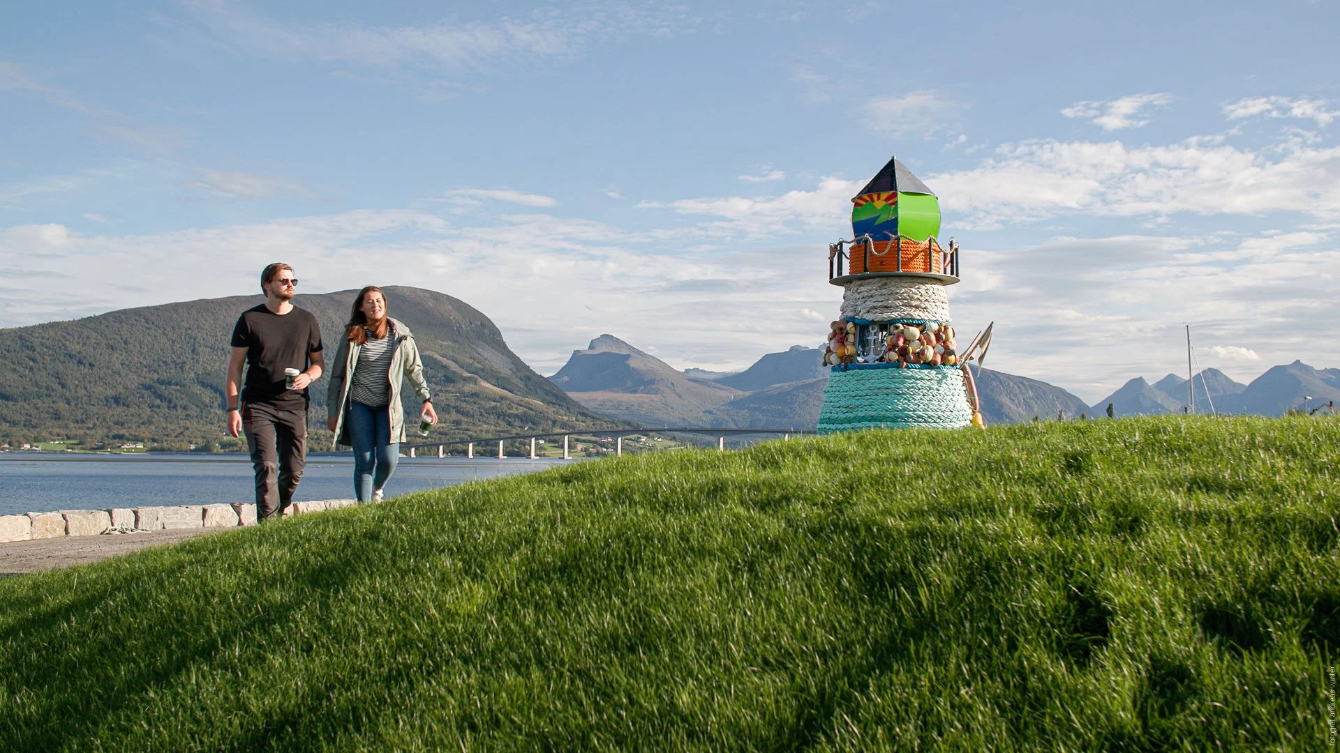 To personer går langs en vannkant, forbi et fyrtårn med røde og rønne detaljer. I bakgrunnen ser man havet og fjellene i Vestnes