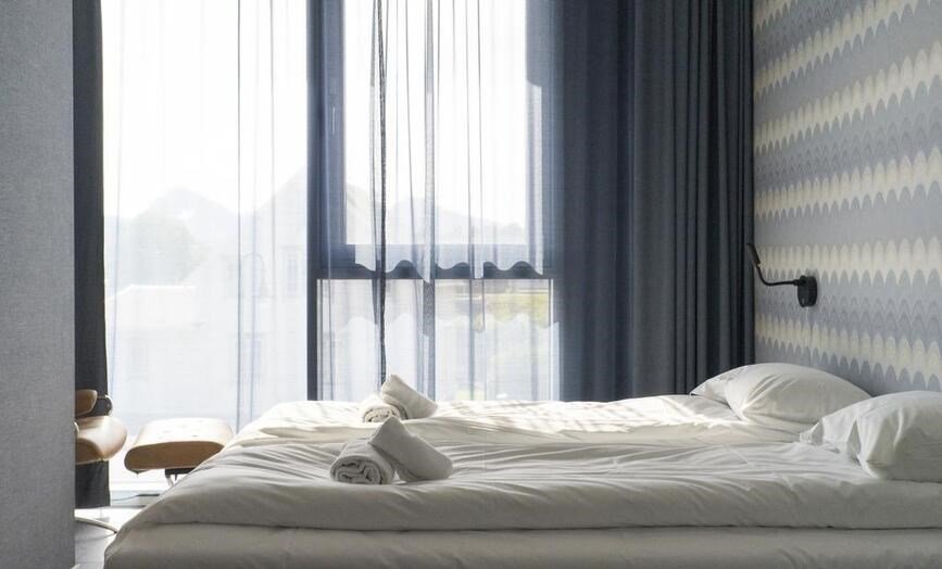 Et rom med seng, håndklær, pute, vindu med gardiner og utsikt utendørs.