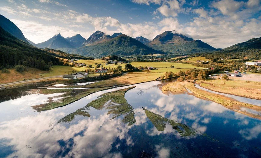 Fjell med skyer, refleksjon av fjell og sky i en vannet, enger og spredte bygninger i et landskap.