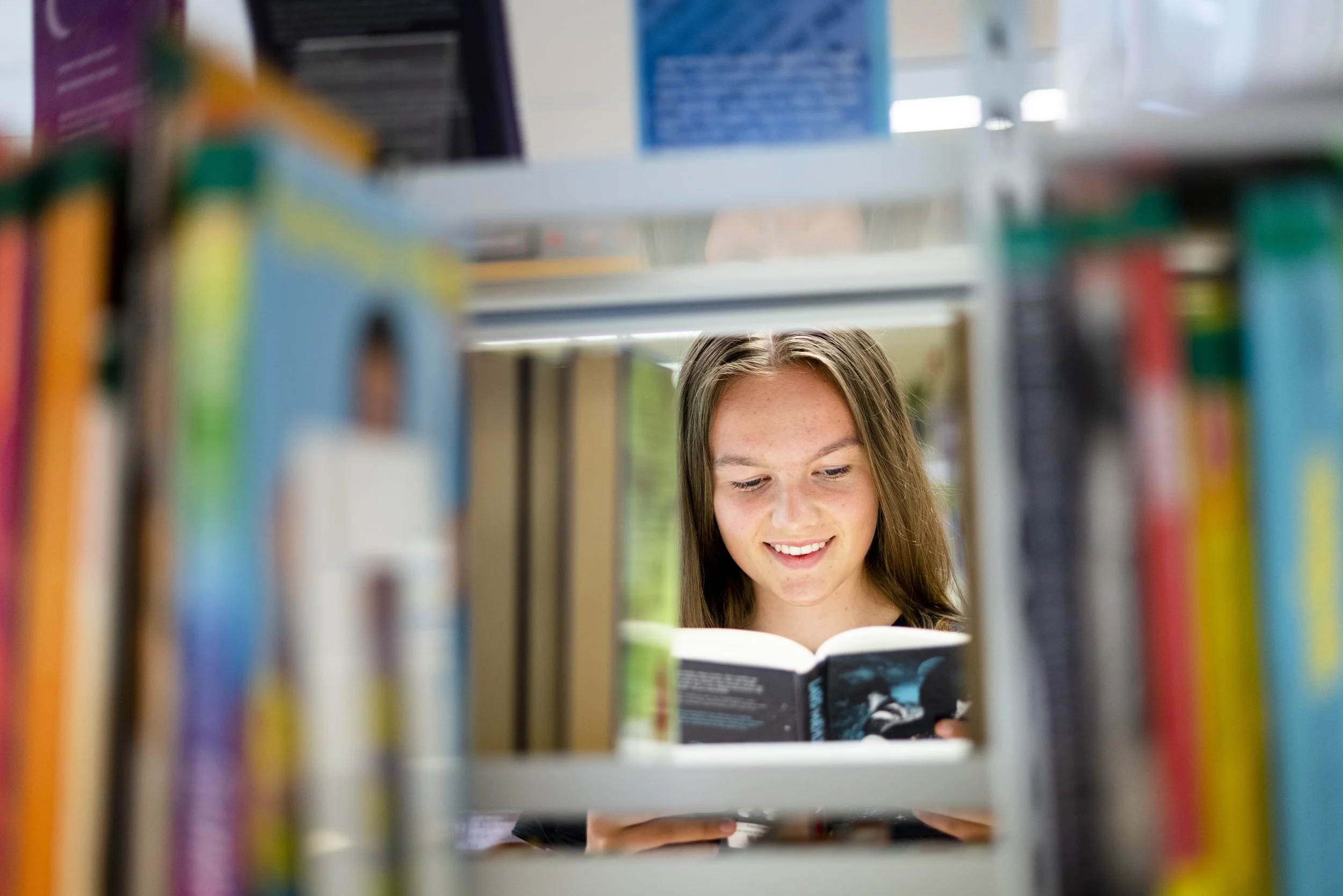 En jente som leser en bok, sett gjennom en hylle på biblioteket.