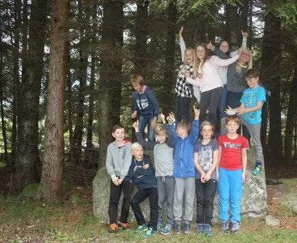 En gruppe barn som står og sitter på og rundt steiner i skogsområde.