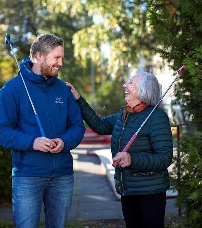 Ei eldre dame og ein yngre mann går tur på ein asfaltert veg, med tre og buskar rundt. Dei ser på kvarandre og kvinna held ei hand på skuldra til mannen