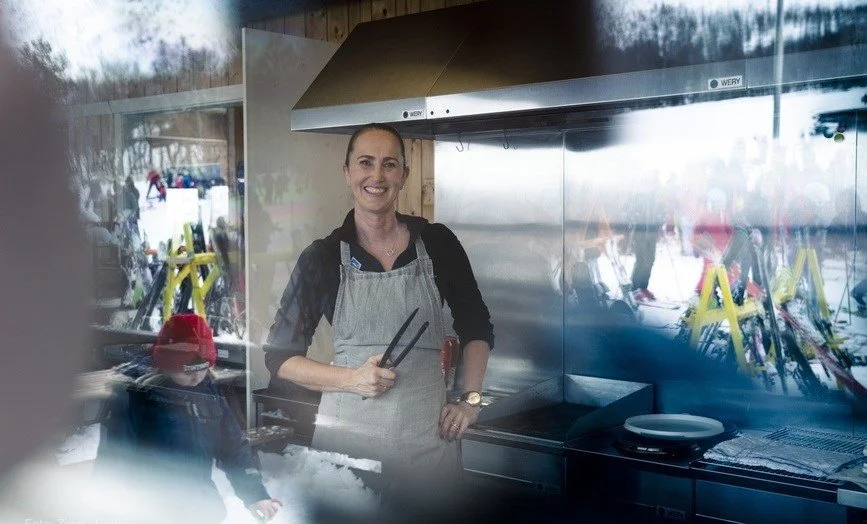 Kvinne som smiler med grillutstyr i et kjøkken. i refleksjonene i vinduet ser man barn og skiutstyr