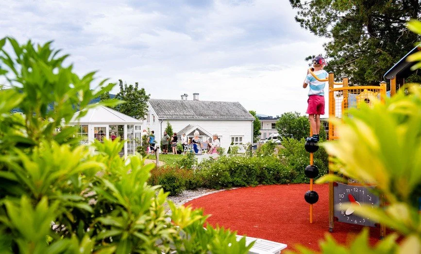 Barn som leker på en utendørs lekeplass. I bakgrunnen ser man grønne busker og plen, et hvitt lysthus, flere mennesker og et hvitt trehus