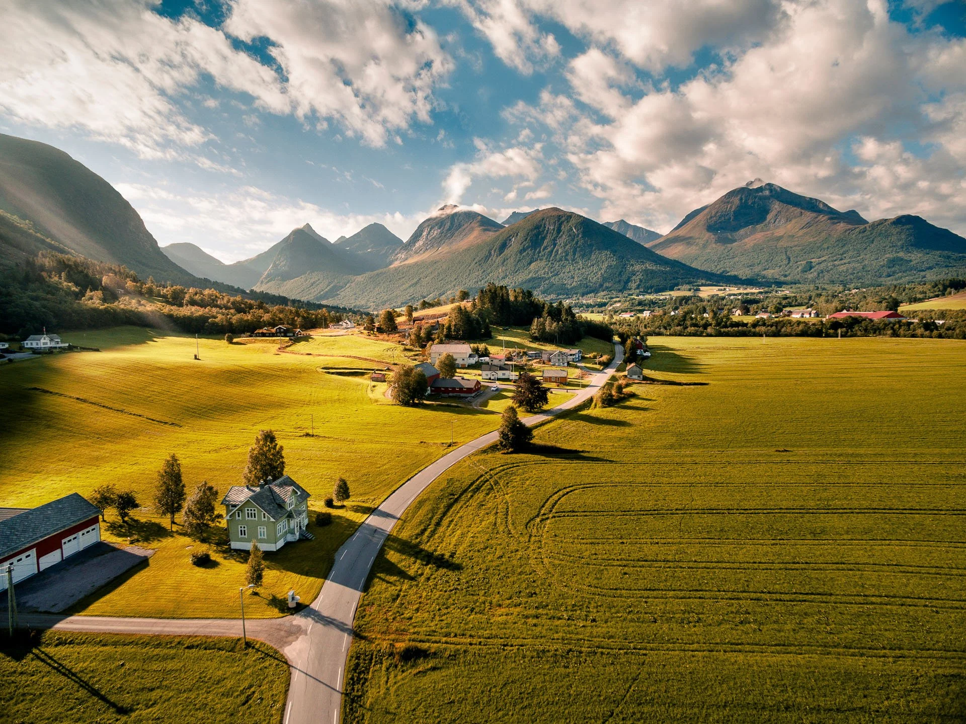 Landskap med grønne åkre, fjell i bakgrunnen og en liten landsby under en skyet himmel.