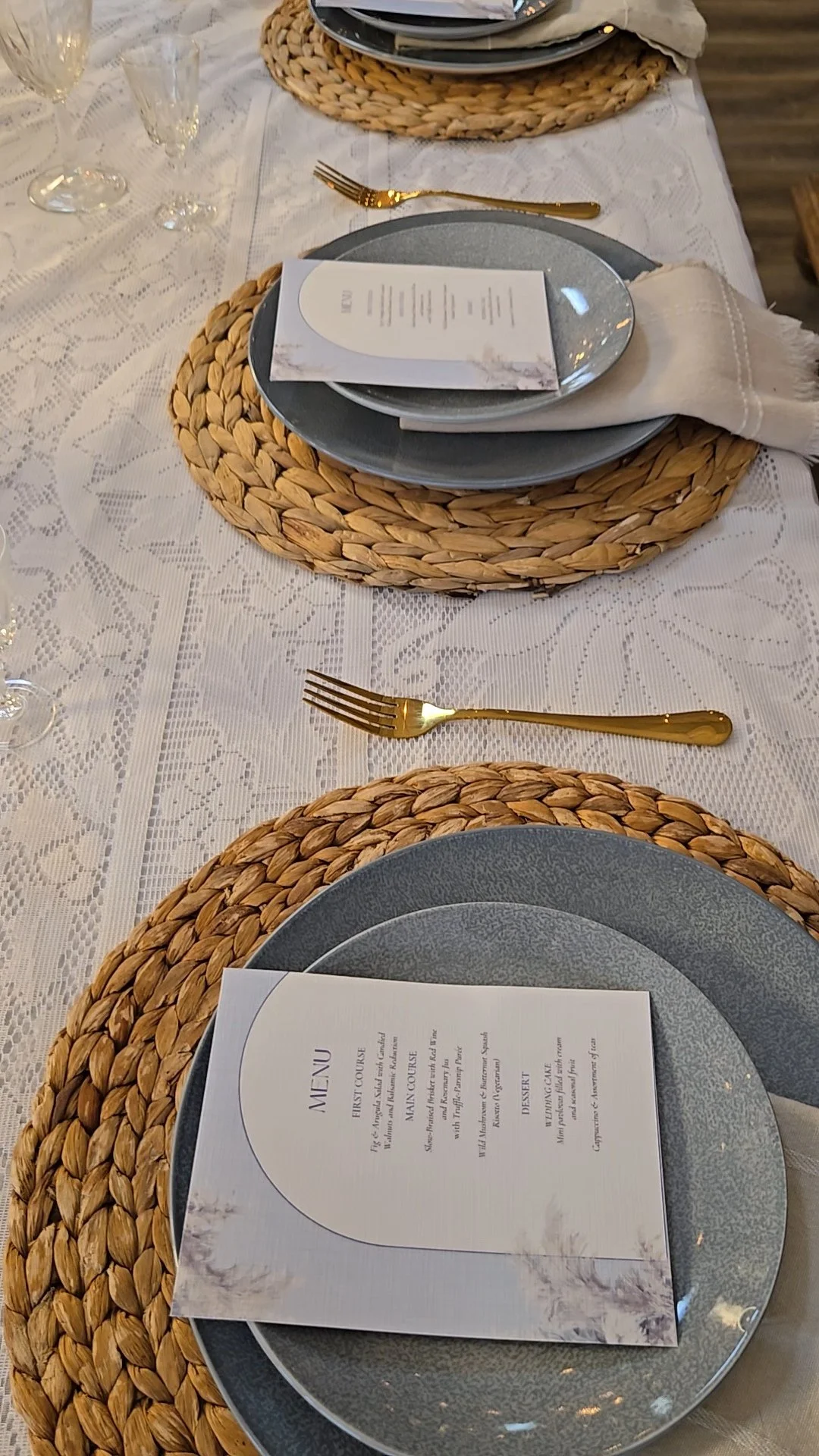 Elegant dining table set with woven placemats, grey plates, gold utensils, a white menu on each setting, and a lace tablecloth.