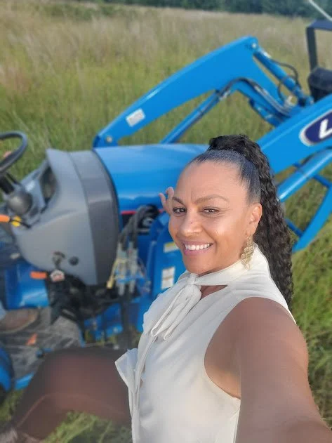 A woman taking a selfie outdoors in a grassy field with a blue tractor in the background.