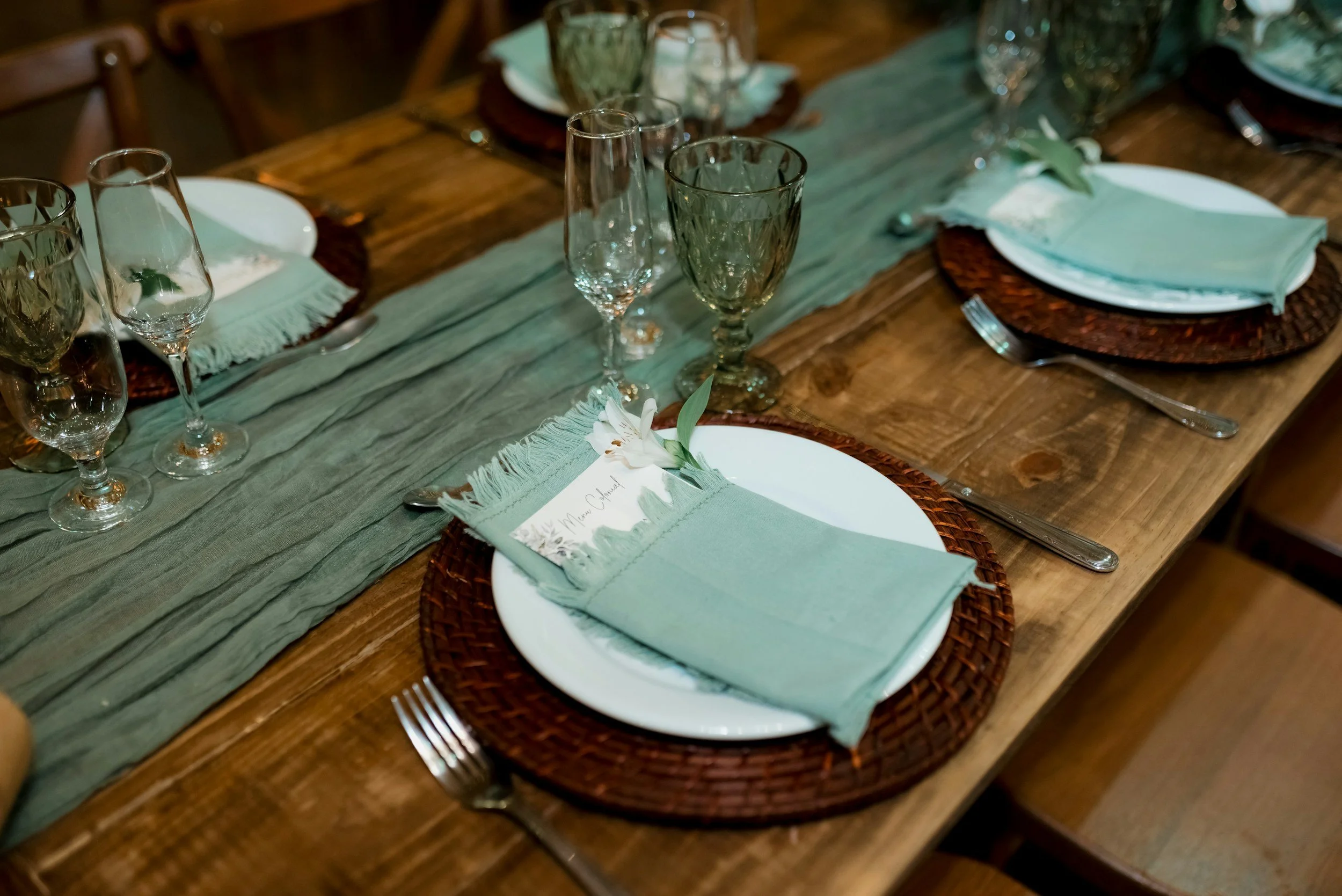 A set dining table with white plates, green napkins, and glassware, decorated with a green table runner and sprigs of greenery and white flowers.
