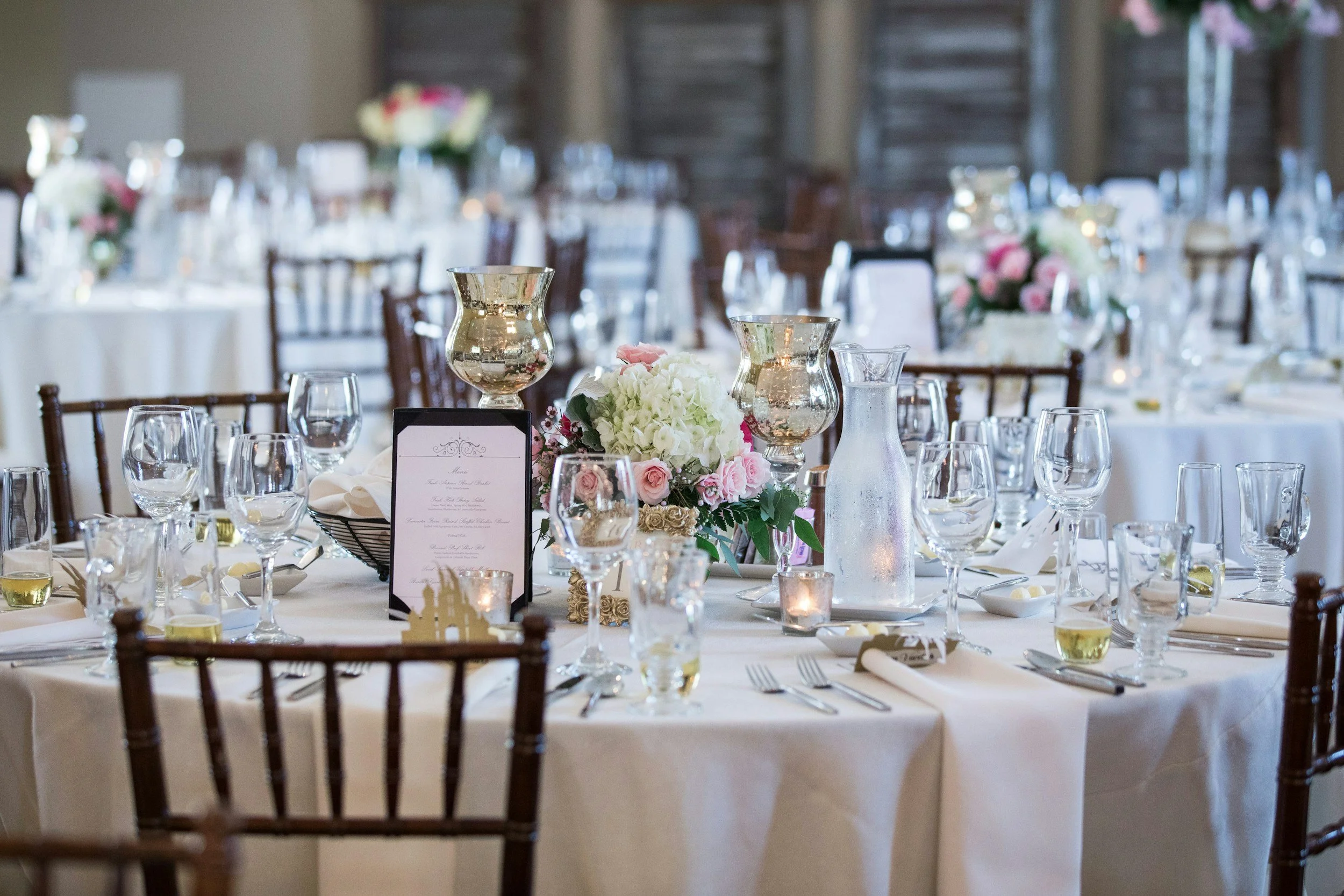 Elegant banquet table decorated with white tablecloths, floral centerpieces with white and pink flowers, candle holders, and glassware, set for a wedding or formal event.