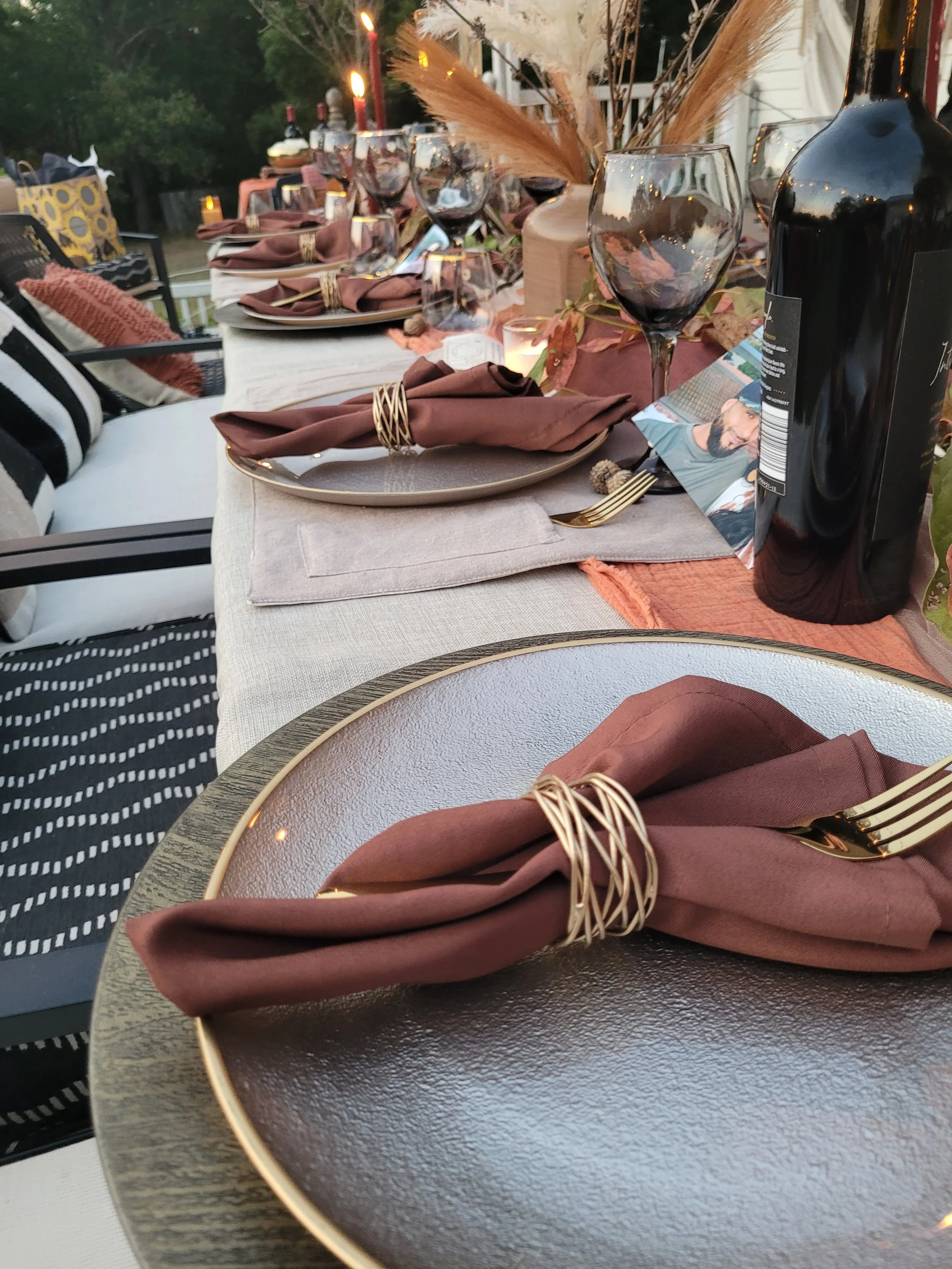 A beautifully set outdoor dining table with dishware, wine glasses, and a bottle of red wine, decorated with candles, pampas grass, and fall leaves for a cozy autumn dinner.