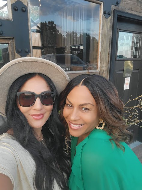 Two women taking a selfie outdoors, smiling, one wearing sunglasses and a hat, and the other with shoulder-length hair and wearing a green top.
